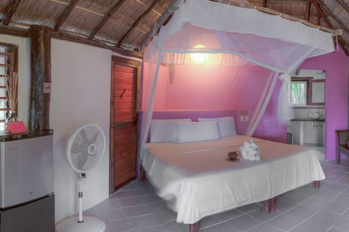 A bedroom with a bed under a canopy, a fan, a small table with a decorative item, and a bathroom visible through an open door.