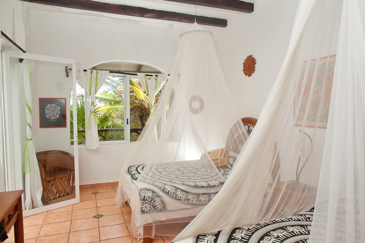 A bedroom with a bed under a canopy, a window with curtains, a wooden chair, and a small table.