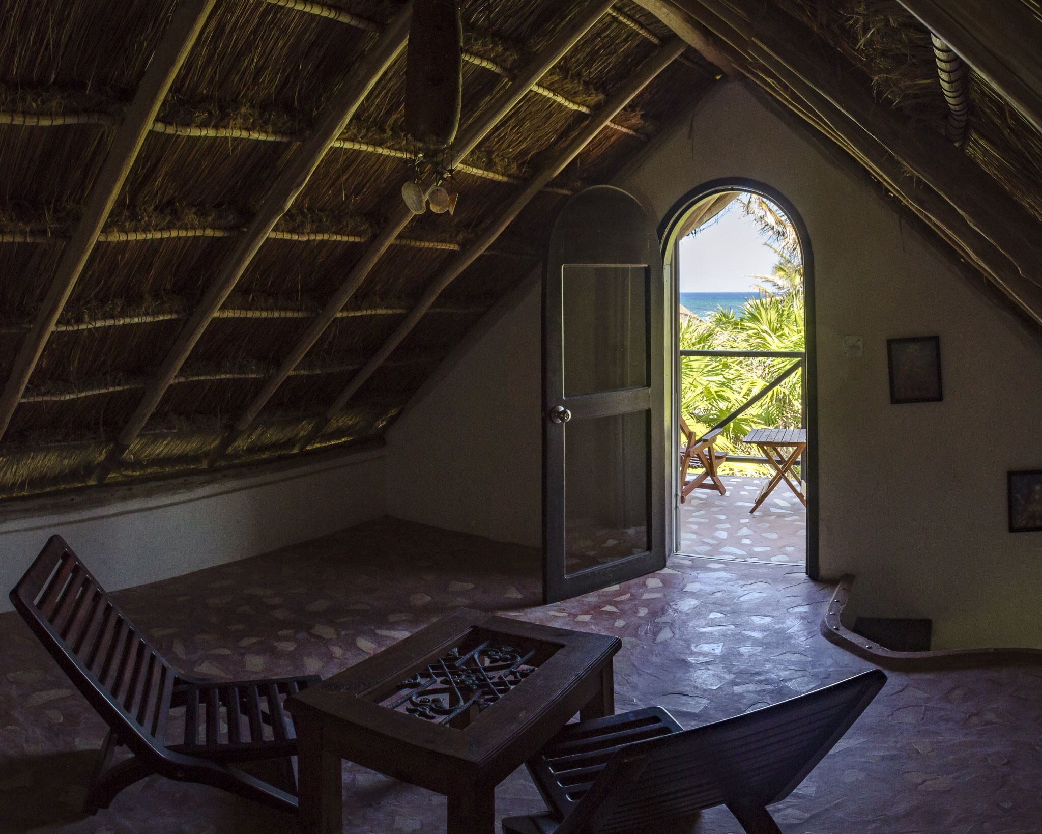 A cozy attic room with a sloped ceiling, a wooden table, two chairs, and a door leading to a balcony with a view of the beach.