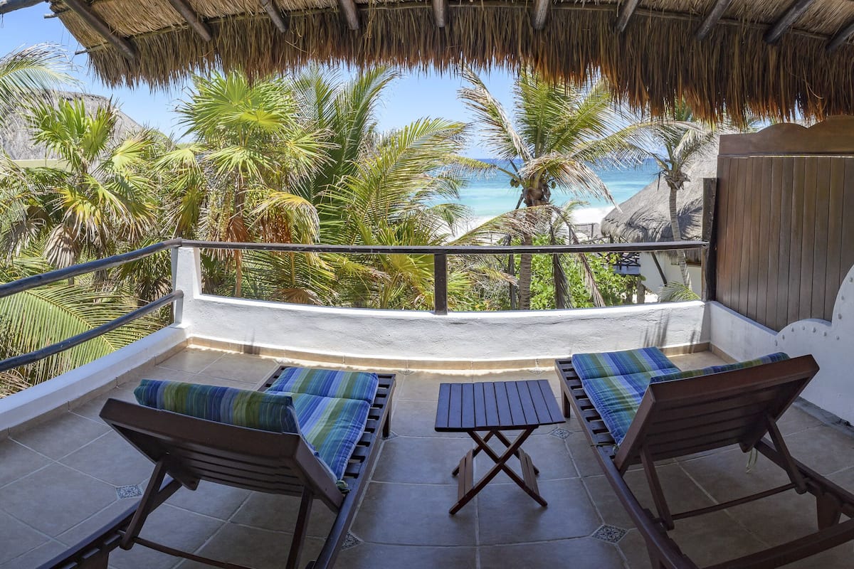 A balcony with two wooden chairs and a small table, overlooking a beach with palm trees and the ocean.