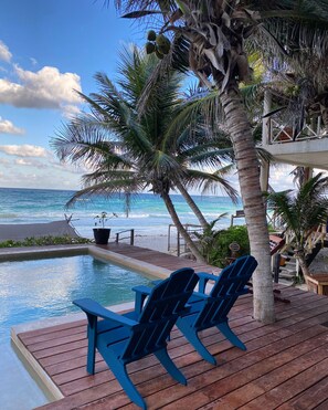A poolside area with a wooden deck, two blue Adirondack chairs, and a view of the ocean.