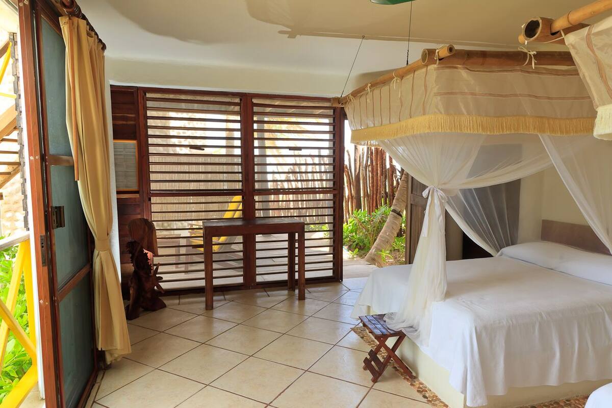 A room with a bed, a small table, and a chair. There is a window with blinds and a curtain.