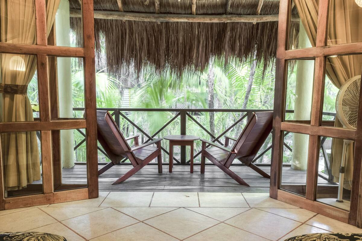 A room with a wooden ceiling, two wooden chairs, a small table, and a view of lush greenery outside.
