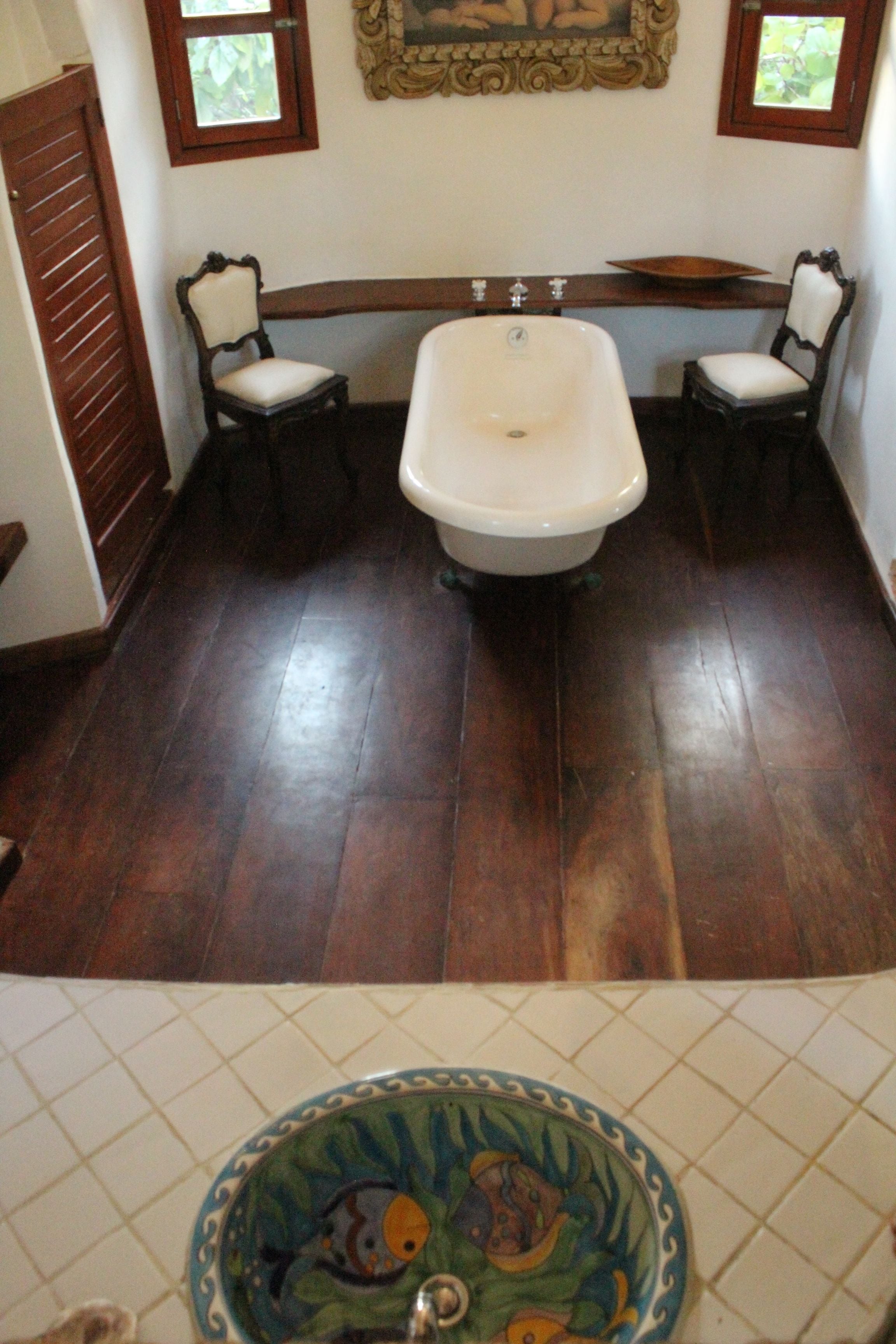 A bathroom with a bathtub, two chairs, a wooden floor, and a tiled sink area.