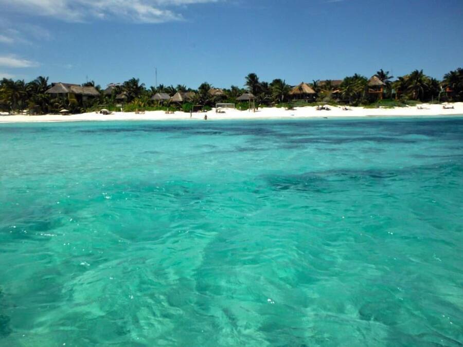 Un resort en la playa con agua turquesa cristalina, arena blanca y cabañas con techo de paja.