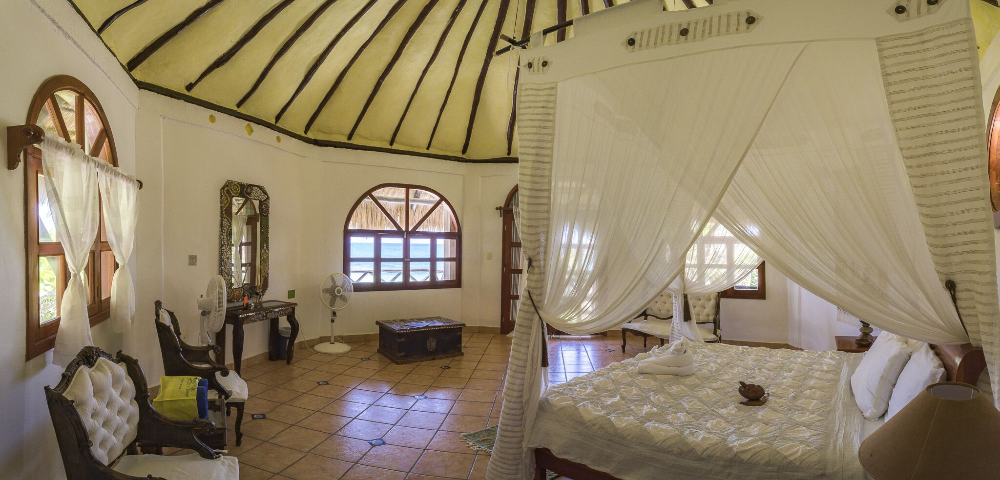 Un dormitorio con cama con dosel, una zona de estar con una silla y una mesa, un espejo y una ventana con vistas.