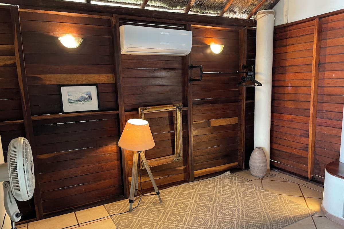 A wooden room with a standing fan, a lamp on a tripod, and a patterned rug on the floor.