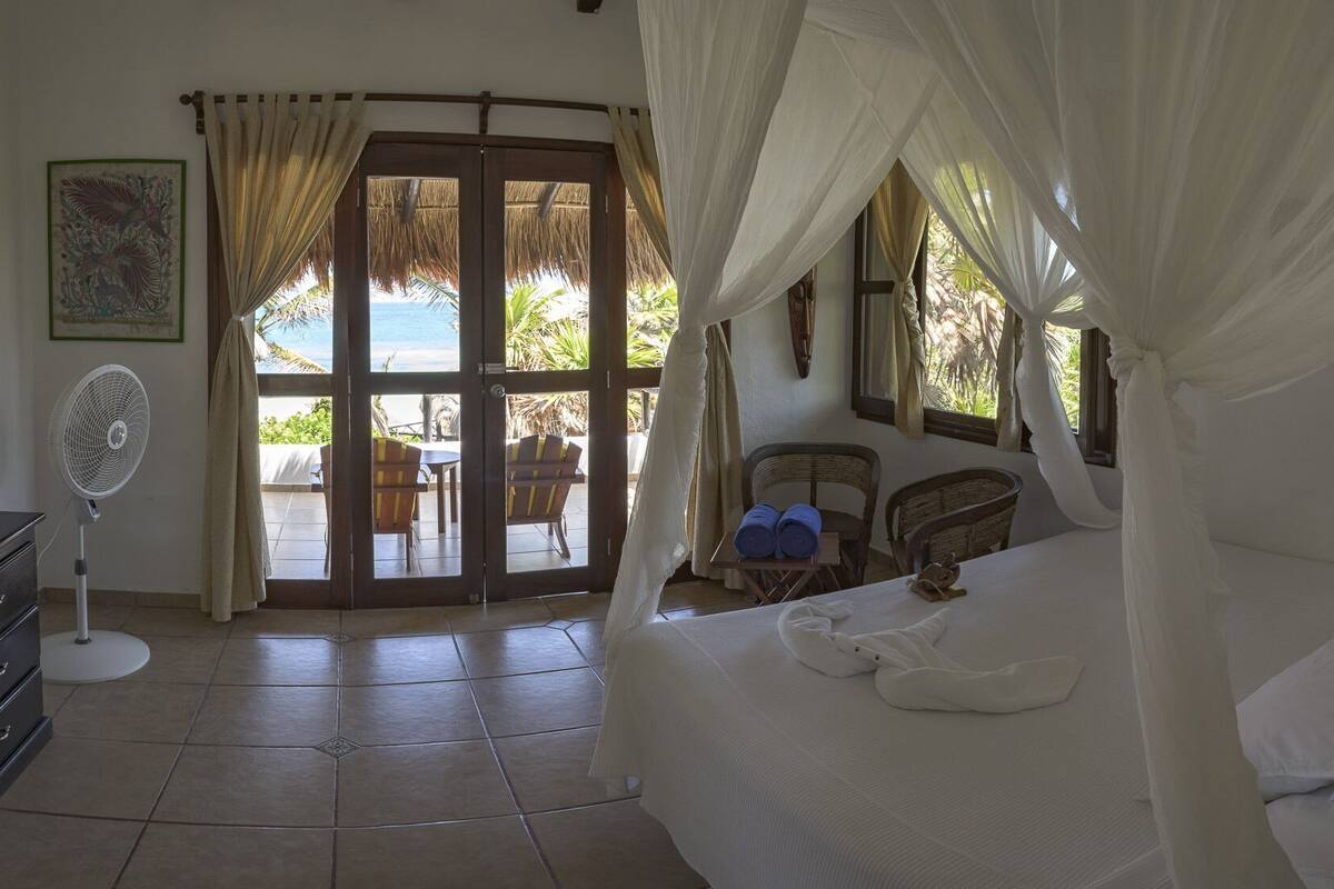 A room with a bed, a fan, a table with chairs, and a view of the beach.