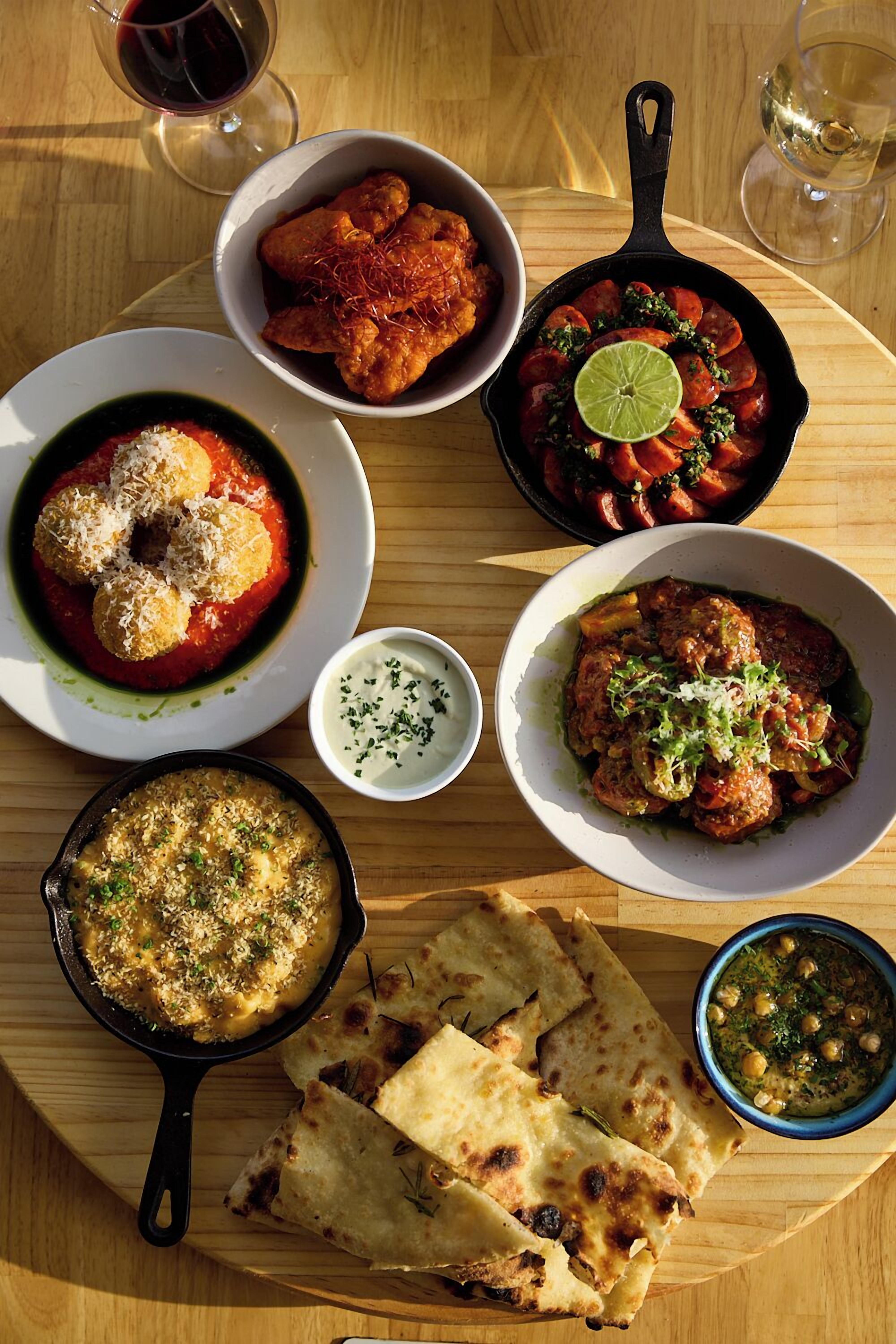 A wooden table with various dishes including a bowl of fried chicken, a skillet with vegetables, and flatbreads.