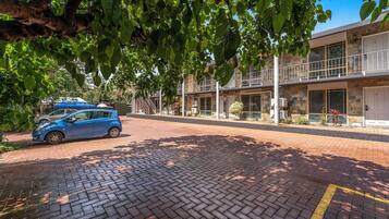 Un coche azul estacionado frente a un edificio con un sendero de ladrillo y un árbol que da sombra.