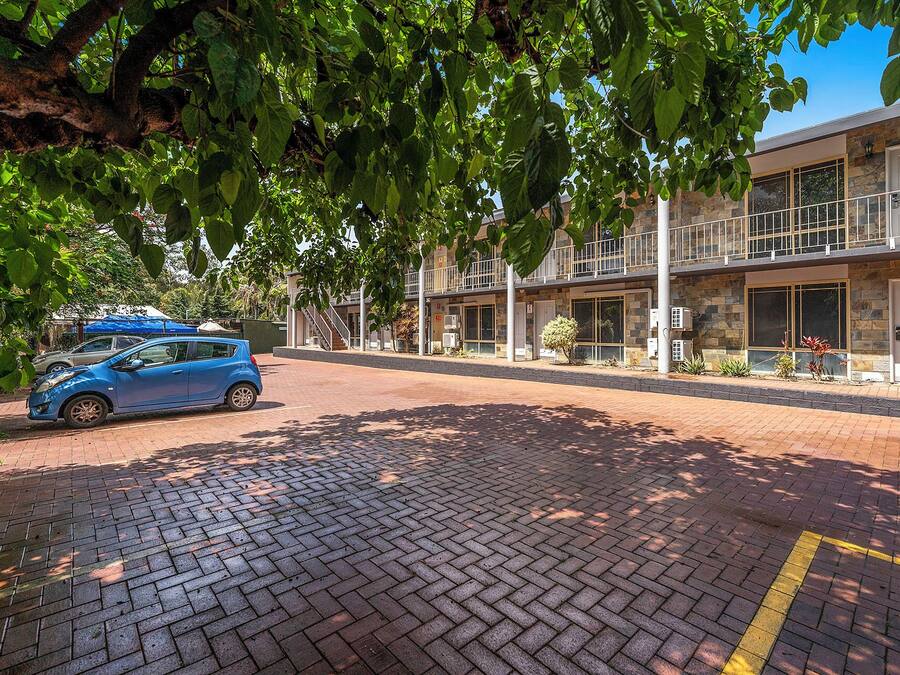 Un coche azul estacionado frente a un edificio con un sendero de ladrillo y un árbol que da sombra.
