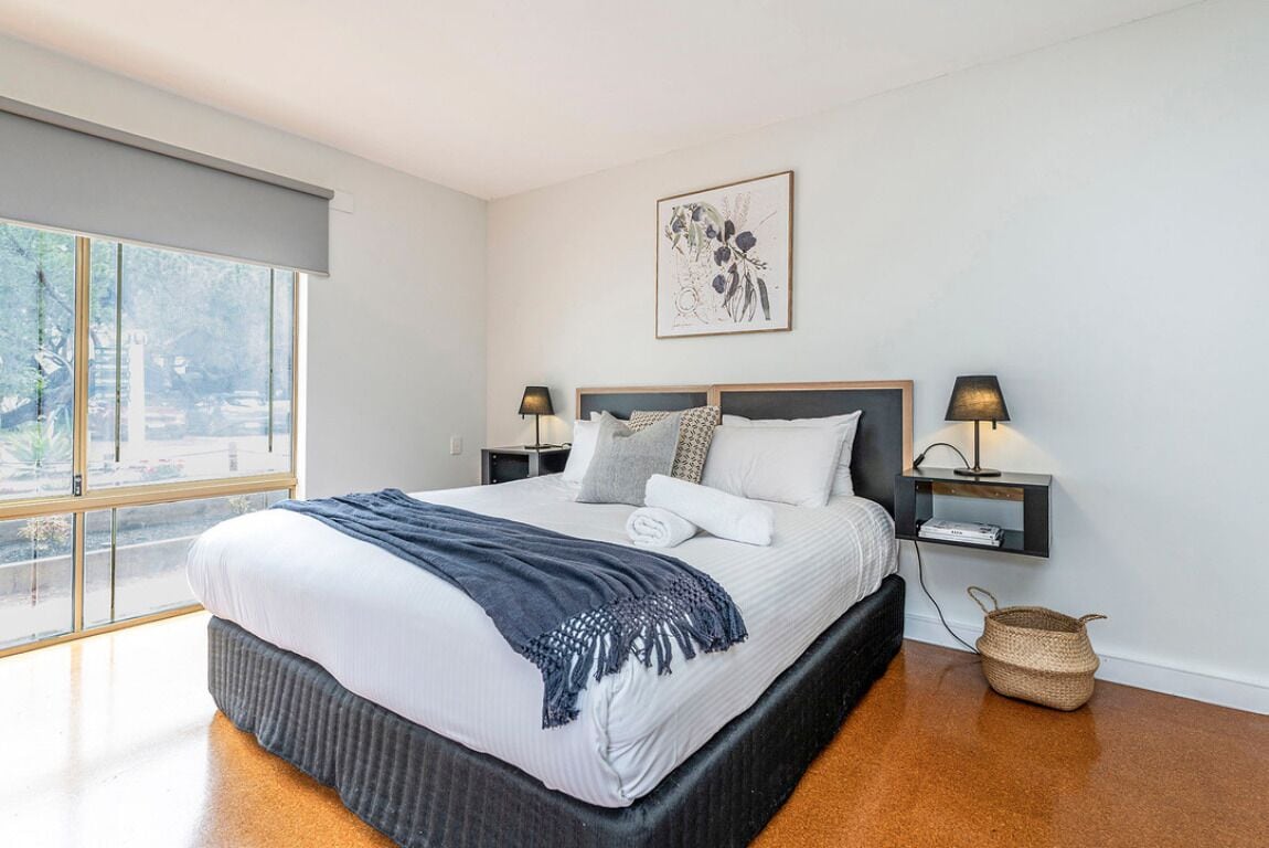 A modern bedroom with a large bed, two bedside tables, a wall-mounted artwork, and a view through a large window.