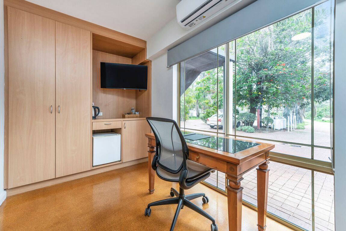 Una oficina moderna con un escritorio de madera, una silla de oficina negra, un televisor de pantalla plana y un amplio ventanal con vistas a árboles y un estacionamiento.