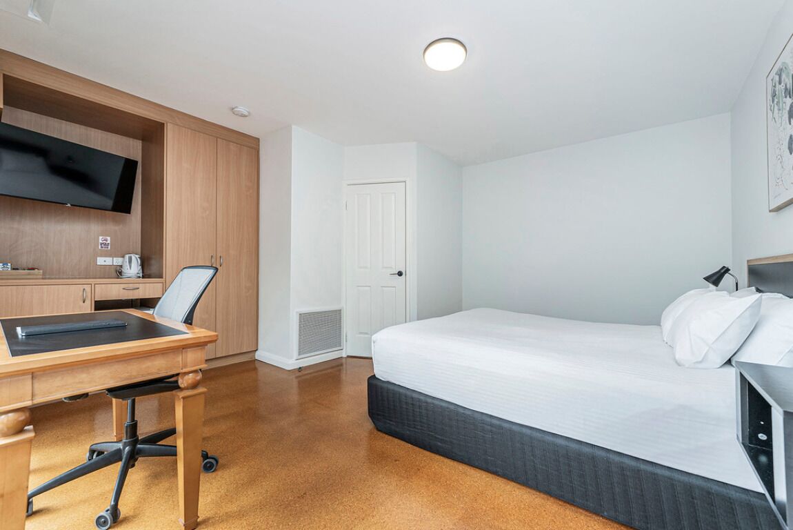 A hotel room with a bed, a desk with a chair, a television mounted on the wall, and a door.
