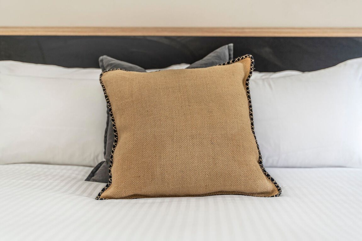 A bed with white bedding and a single beige pillow with a black lace trim.