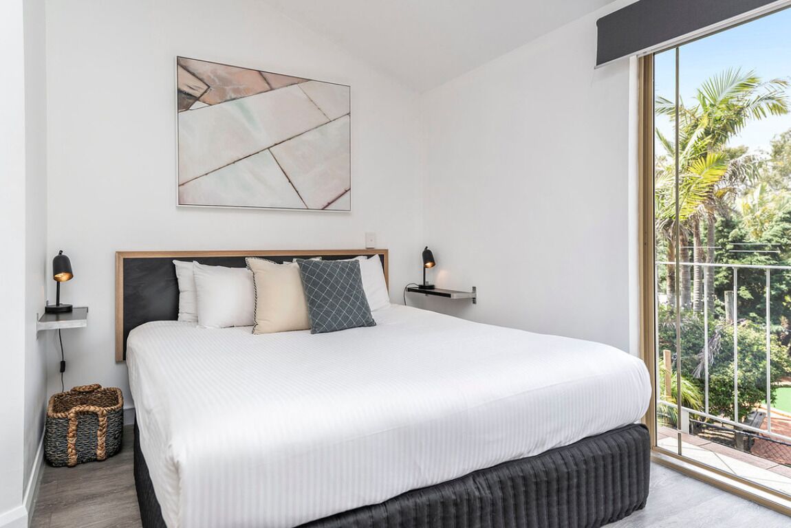 Een slaapkamer met een groot bed, een hoofdbord, een abstract schilderij aan de muur, nachtlampjes en uitzicht op palmbomen door een groot raam.