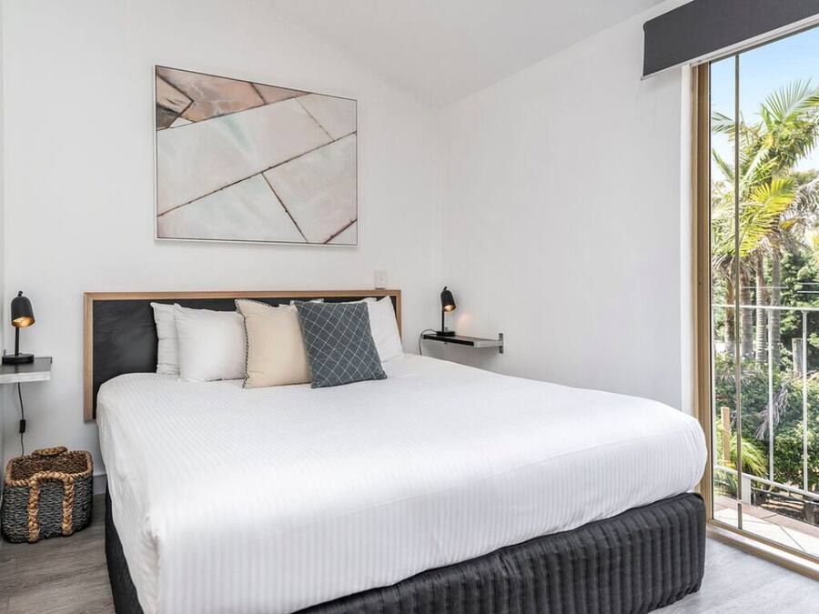 Ein Schlafzimmer mit einem großen Bett, einem Kopfteil, einem an der Wand befestigten abstrakten Gemälde, Nachttischlampen und Blick auf Palmen durch ein großes Fenster.