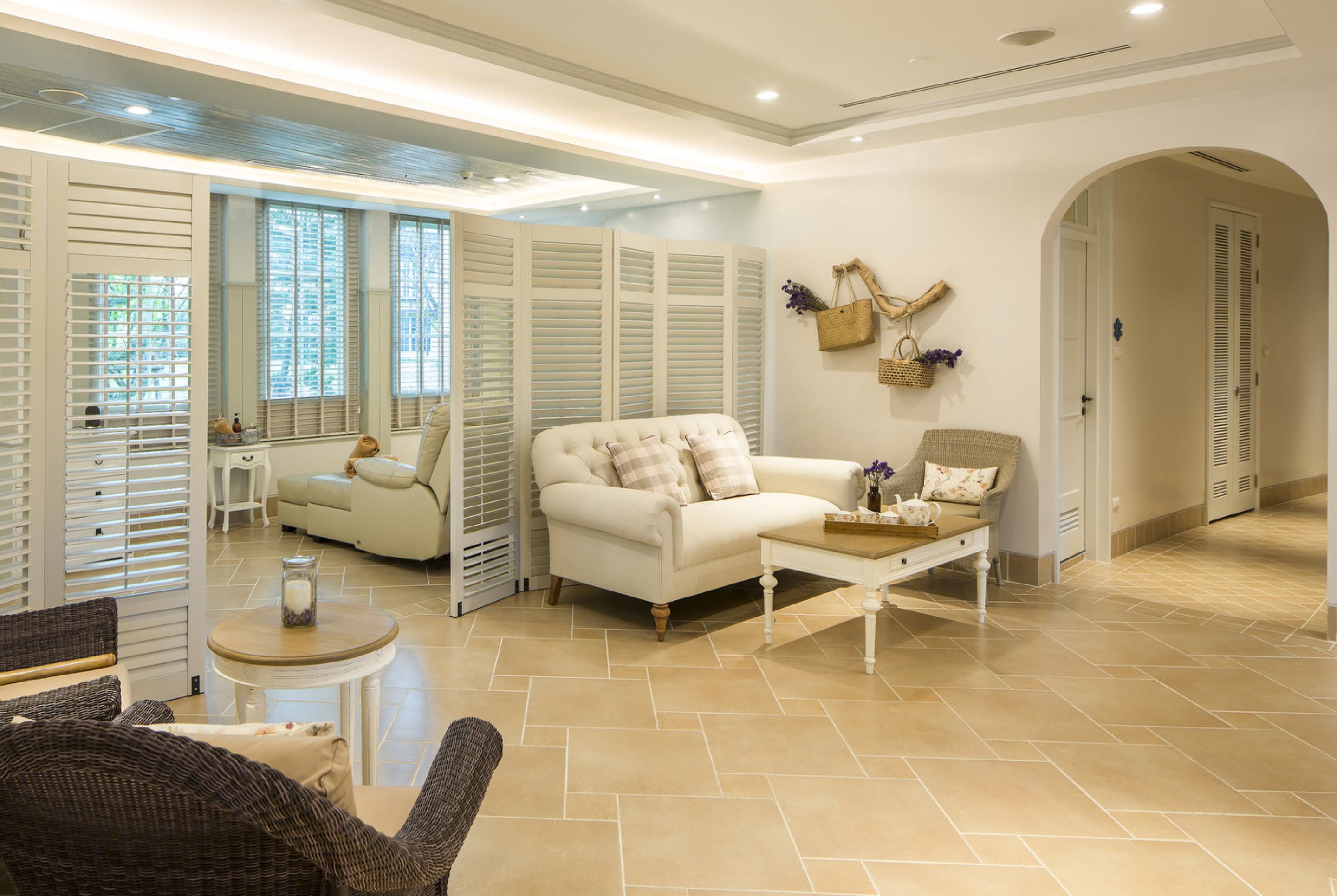 A spacious living room with a white sofa, a coffee table, and wicker chairs. There are large windows with blinds, a white door, and a tiled floor.
