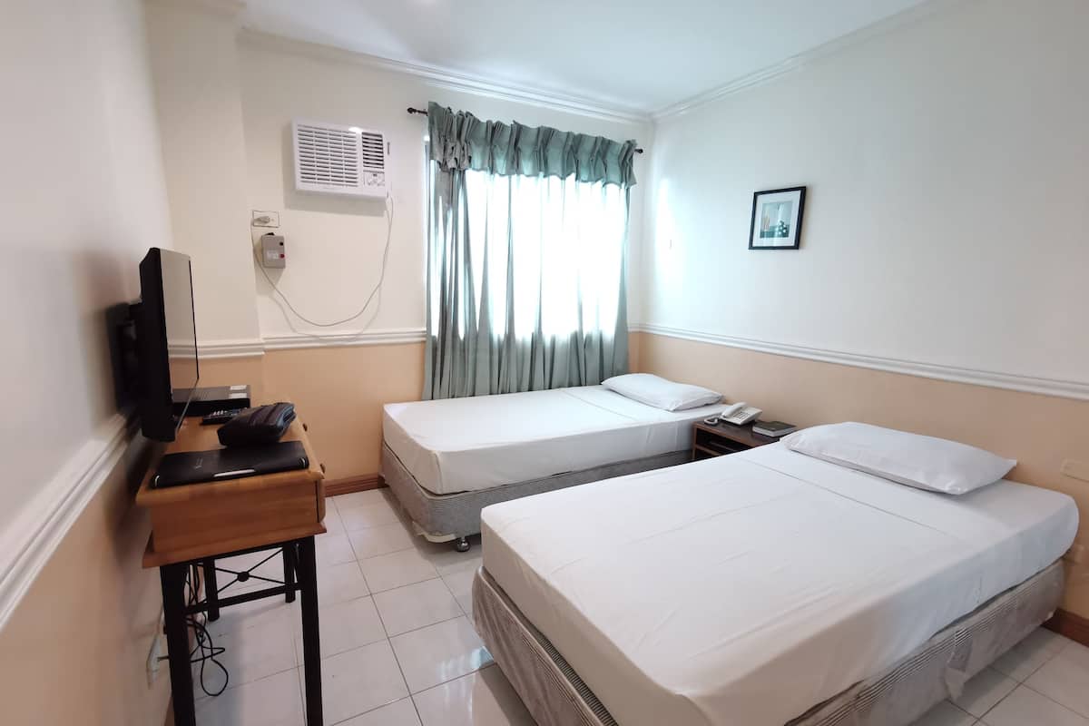 A hotel room with two single beds, a desk with a computer and phone, an air conditioning unit, and a window with curtains.