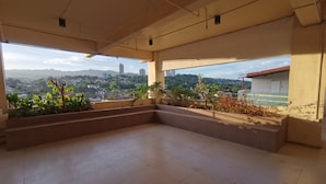 Ein geräumiger Balkon mit Fliesenboden und Topfpflanzen, mit Blick auf eine Stadtlandschaft mit Gebäuden und Hügeln.
