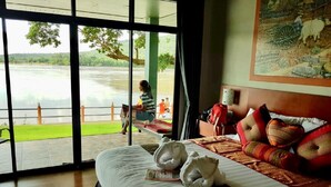 A hotel room with a large window offering a view of a body of water and greenery. There is a bed with pillows and a blanket, and a suitcase on the bed. - Khong Chiam Orchid Riverside Resort (Khong Chiam)