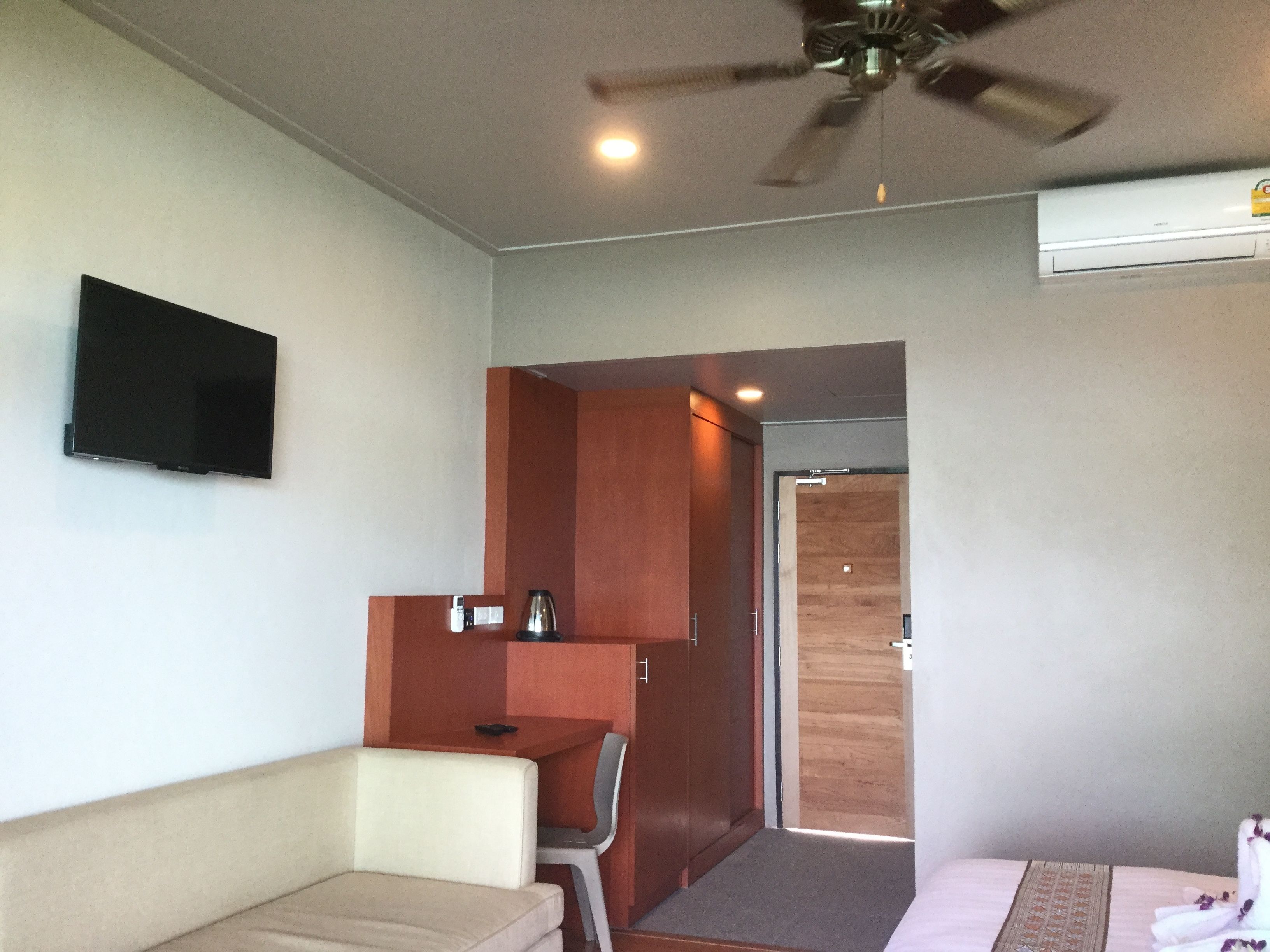 A compact hotel room with a bed, a small desk, a chair, a ceiling fan, and a TV mounted on the wall.