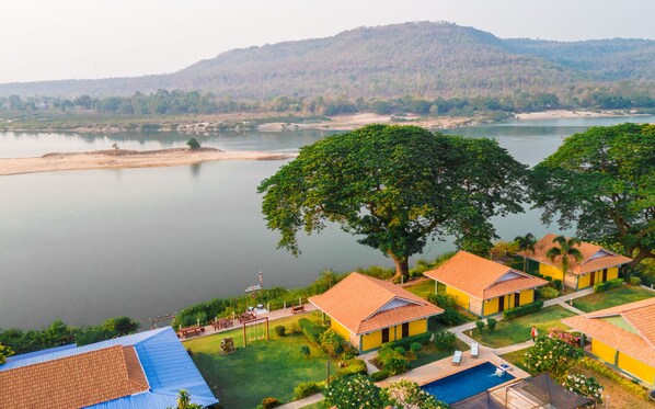 A resort with multiple yellow buildings, a swimming pool, and a view of a calm lake with a forested hill in the background. - Khong Chiam Orchid Riverside Resort (Khong Chiam)