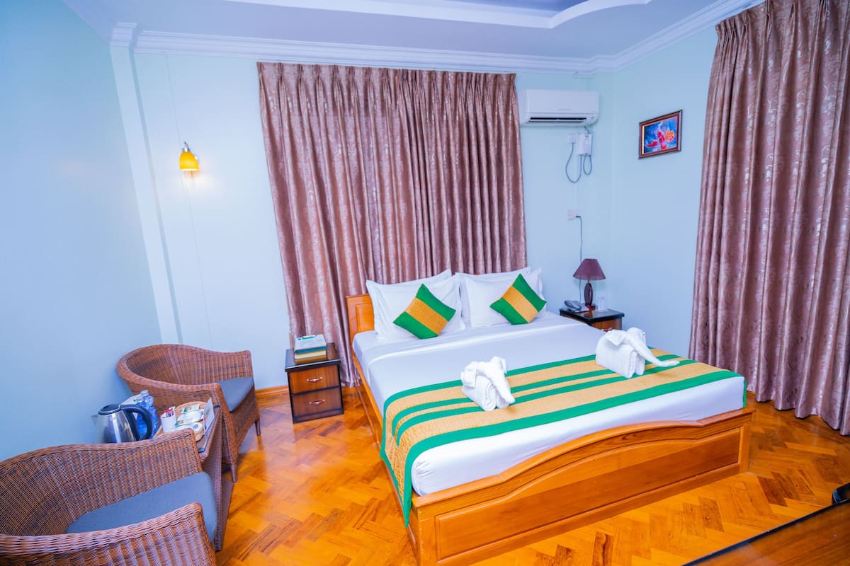 A hotel room with a wooden bed, two wicker chairs, a nightstand, and a wall-mounted air conditioner.