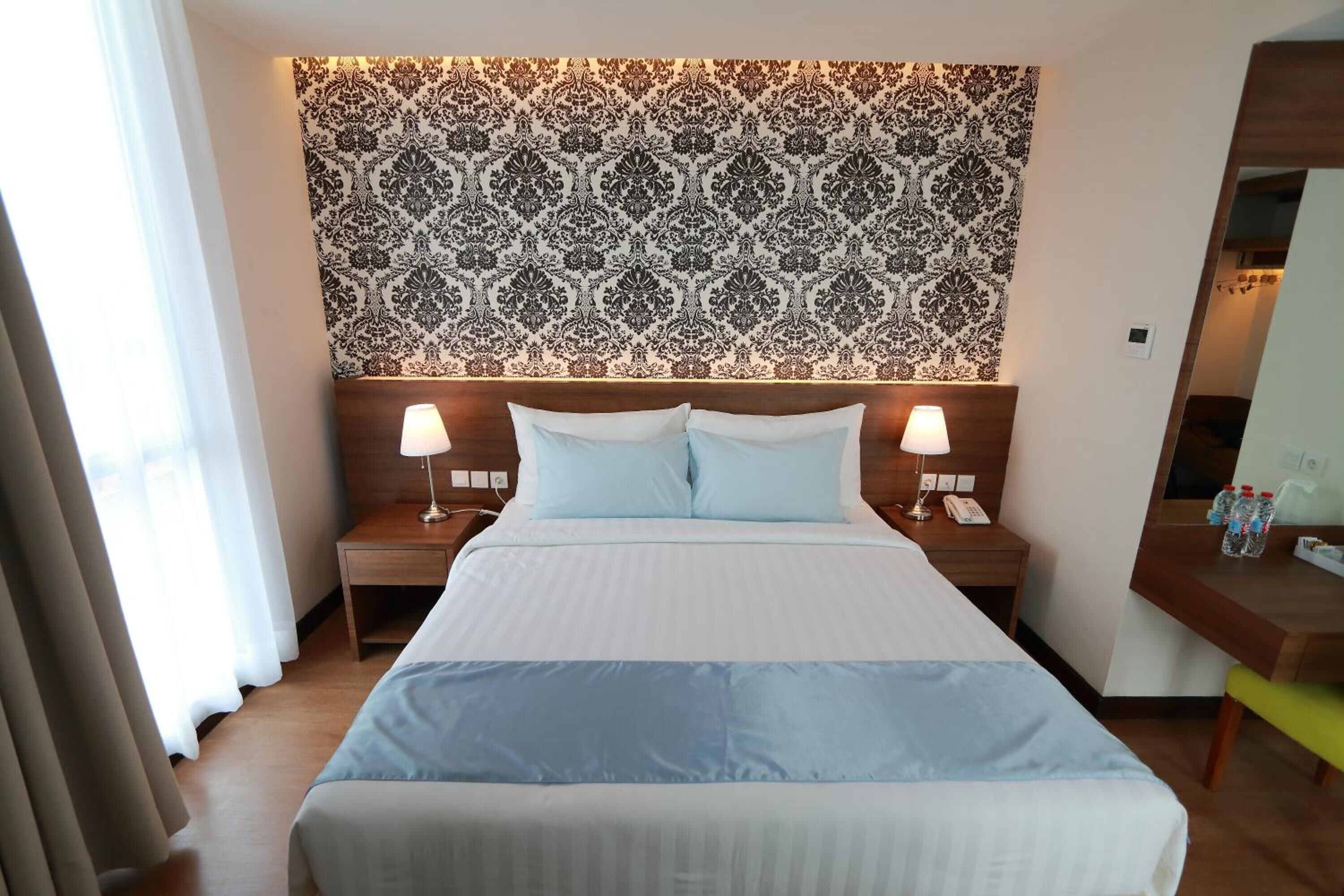 A hotel room with a large bed, two bedside tables with lamps, a patterned headboard, and a small desk with a mirror and water bottles.