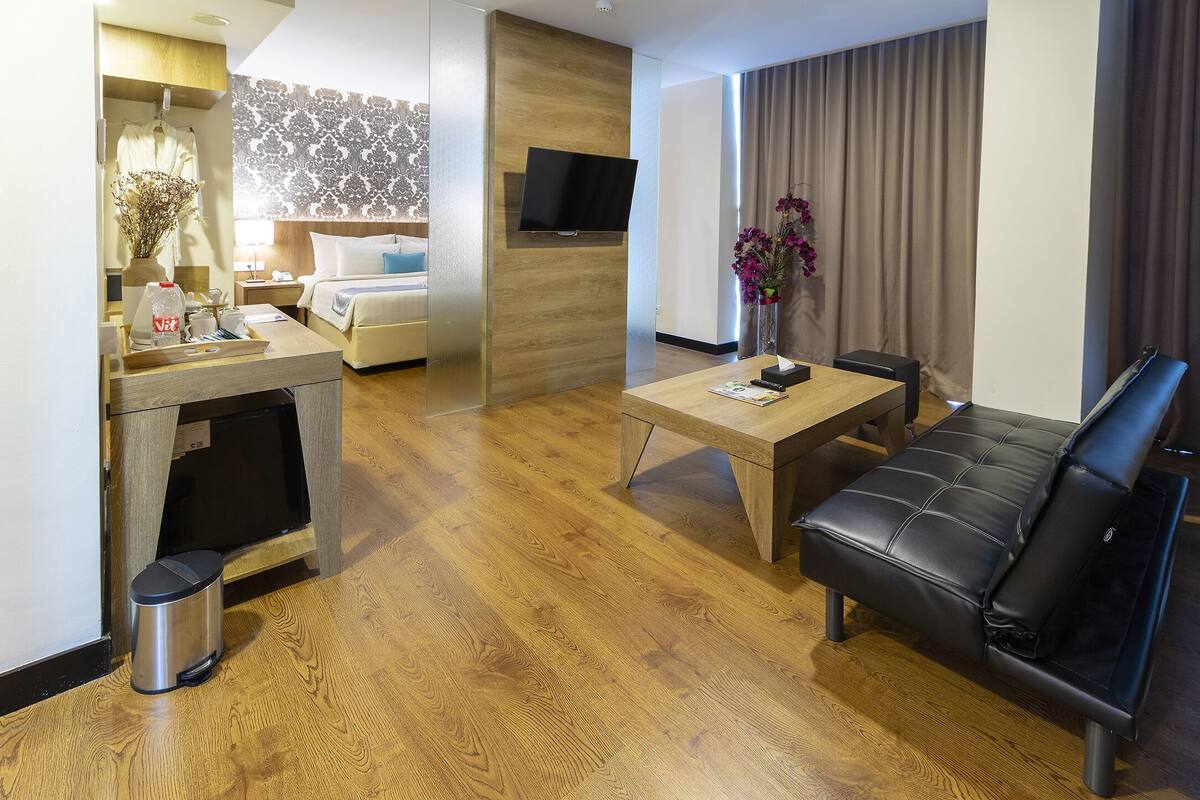 A modern hotel room with a wooden floor, a black leather sofa, a wooden coffee table, a TV mounted on the wall, and a bed with a patterned headboard.