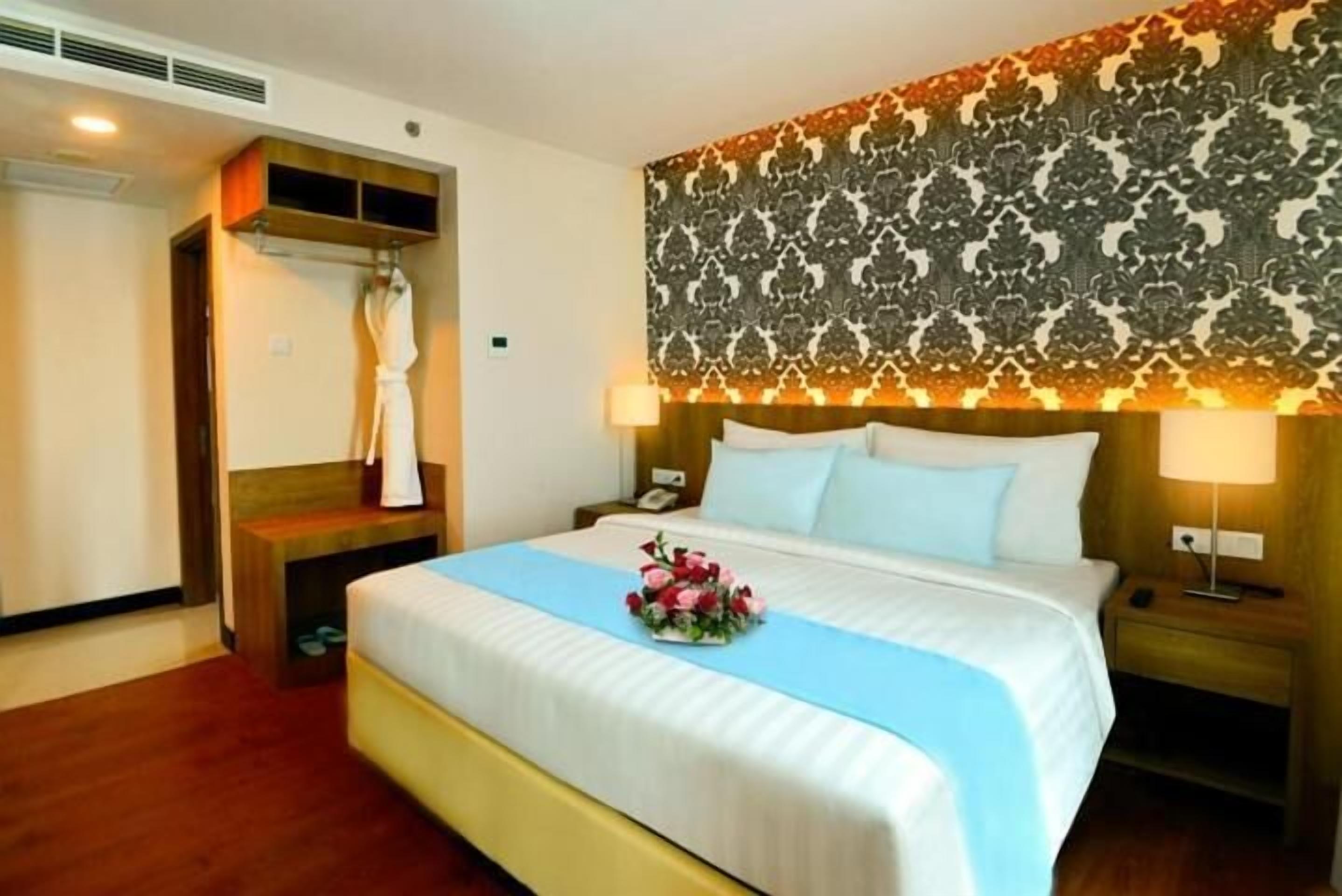 A hotel room with a large bed, a wooden headboard, a patterned wall, and a bedside table with a lamp.