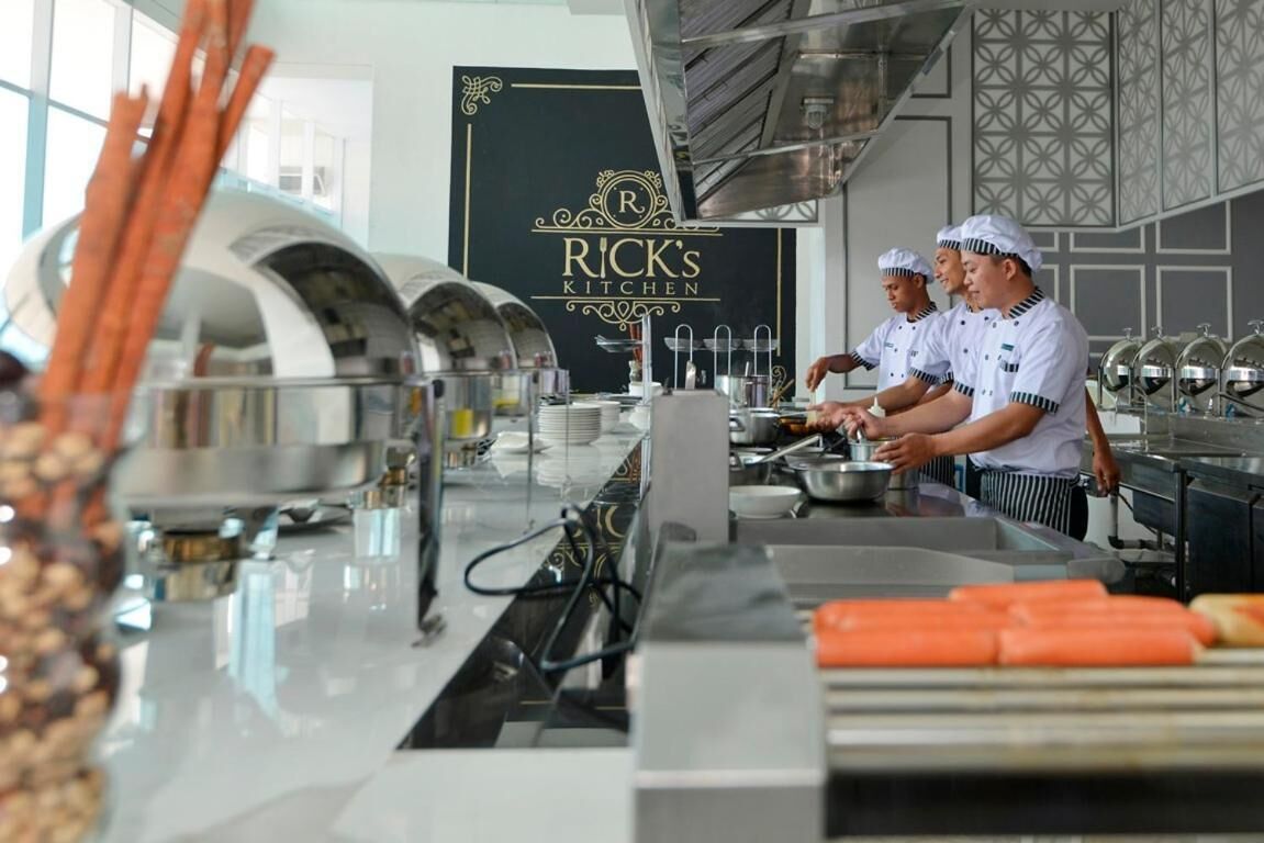 A professional kitchen with chefs preparing food, stainless steel equipment, and a branded sign for Rick's Kitchen.