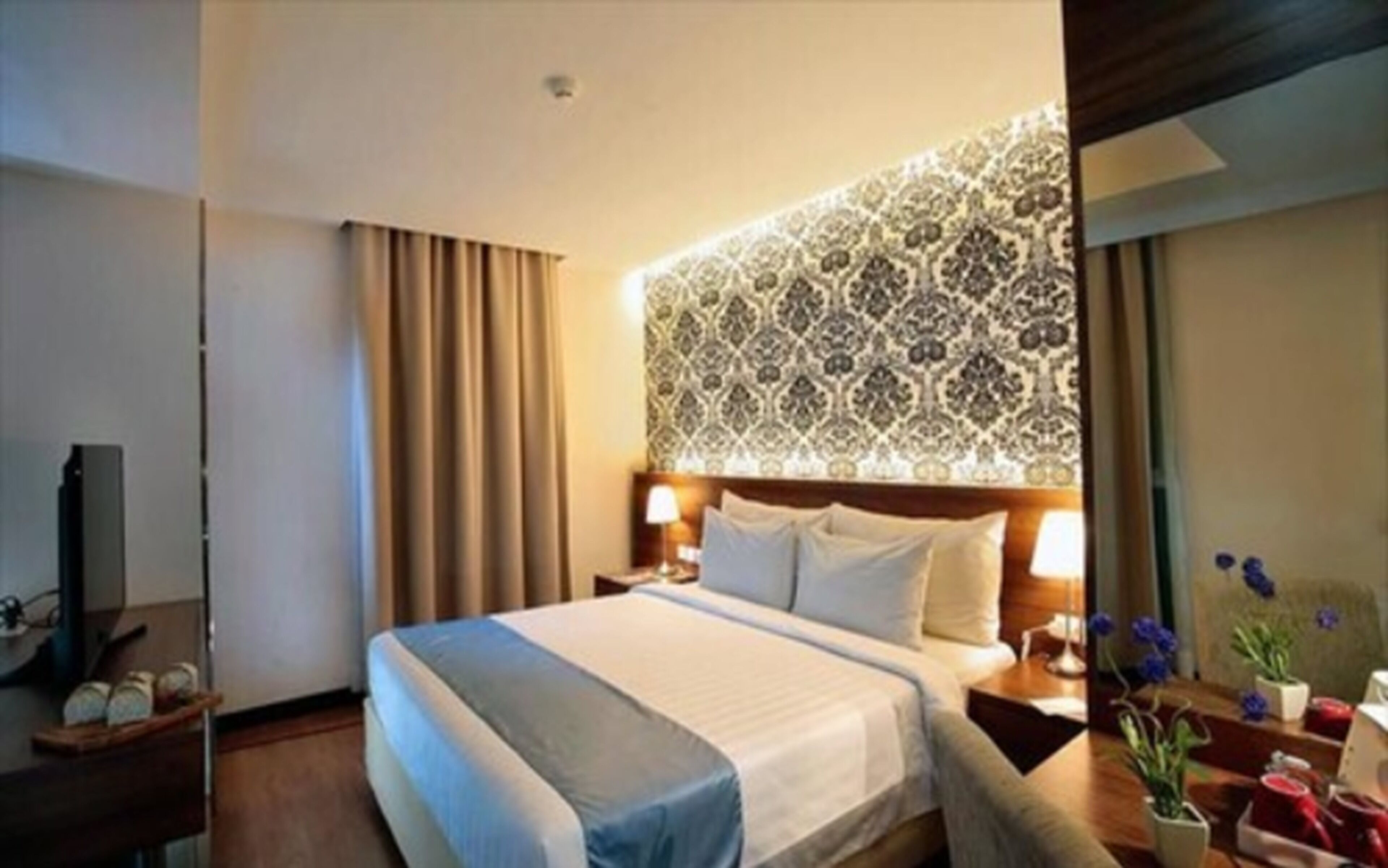A hotel room with a large bed, a headboard with a patterned design, a bedside table with a lamp, and a nightstand with a vase of flowers.