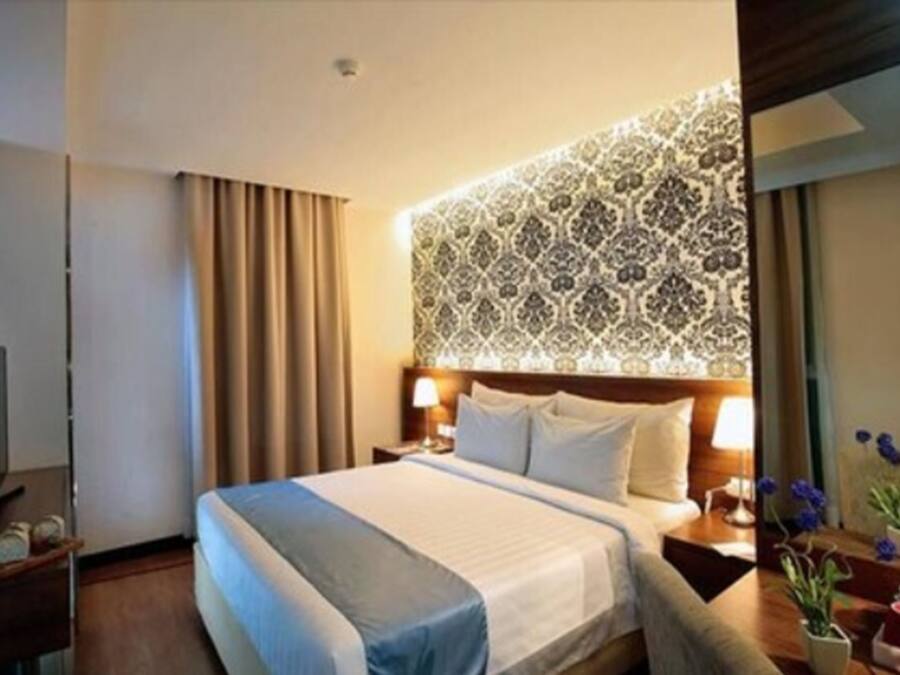 A hotel room with a large bed, a headboard with a patterned design, a bedside table with a lamp, and a nightstand with a vase of flowers.