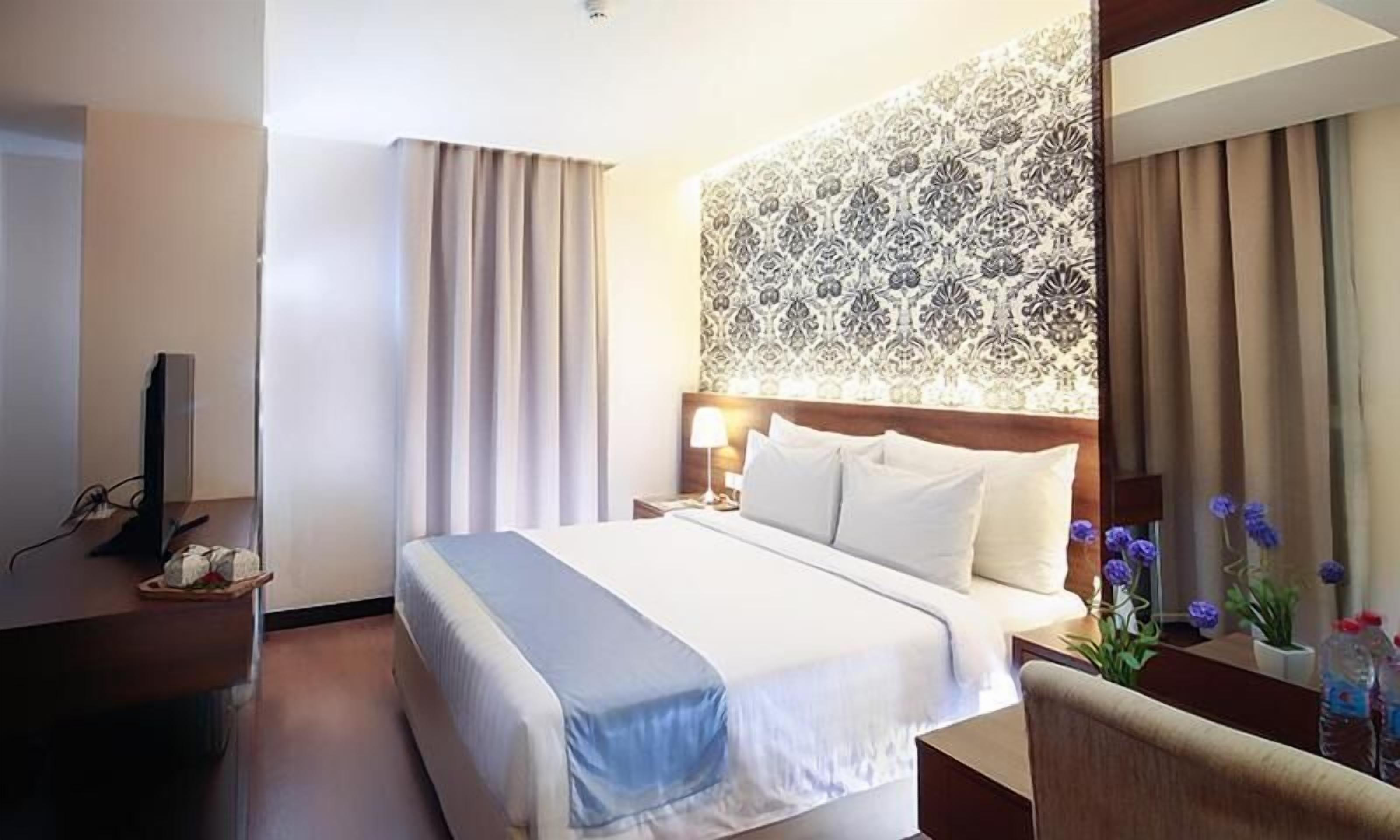 Una habitación de hotel con una cama grande, una mesita de noche con lámpara, un televisor sobre un mueble bajo y un panel decorativo en la pared.