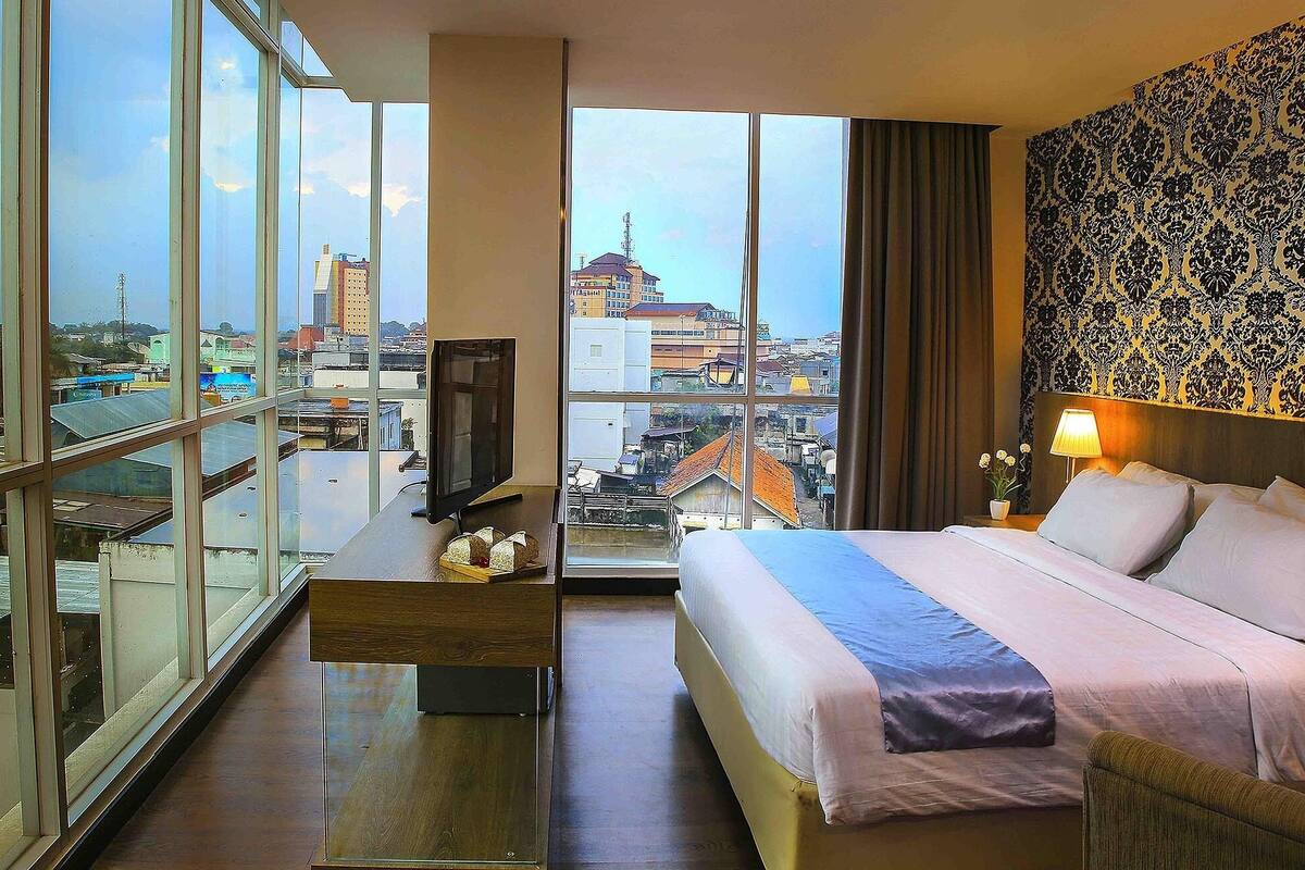 A hotel room with a large window, a bed, a desk, and a television.