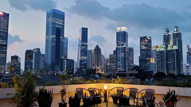 A city skyline with modern skyscrapers, a rooftop terrace with chairs and plants, and a clear sky. - Topaz Residence (Jakarta)