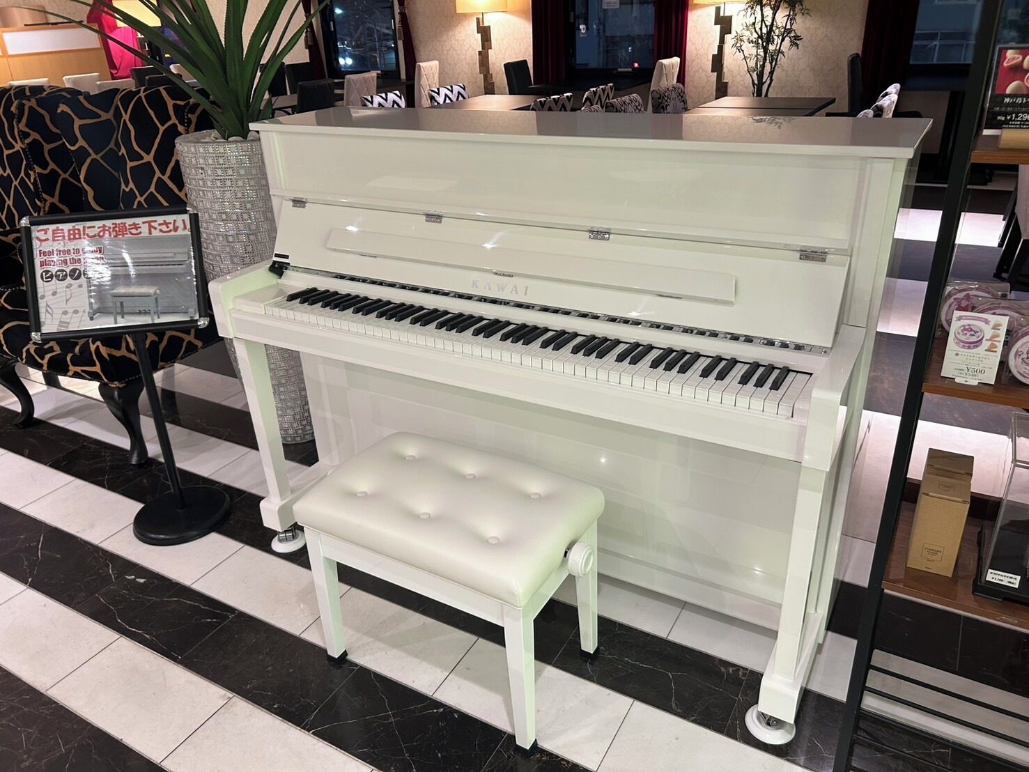 Un piano et un banc dans un espace d’accueil, avec un canapé à motifs et une plante en pot en arrière-plan.