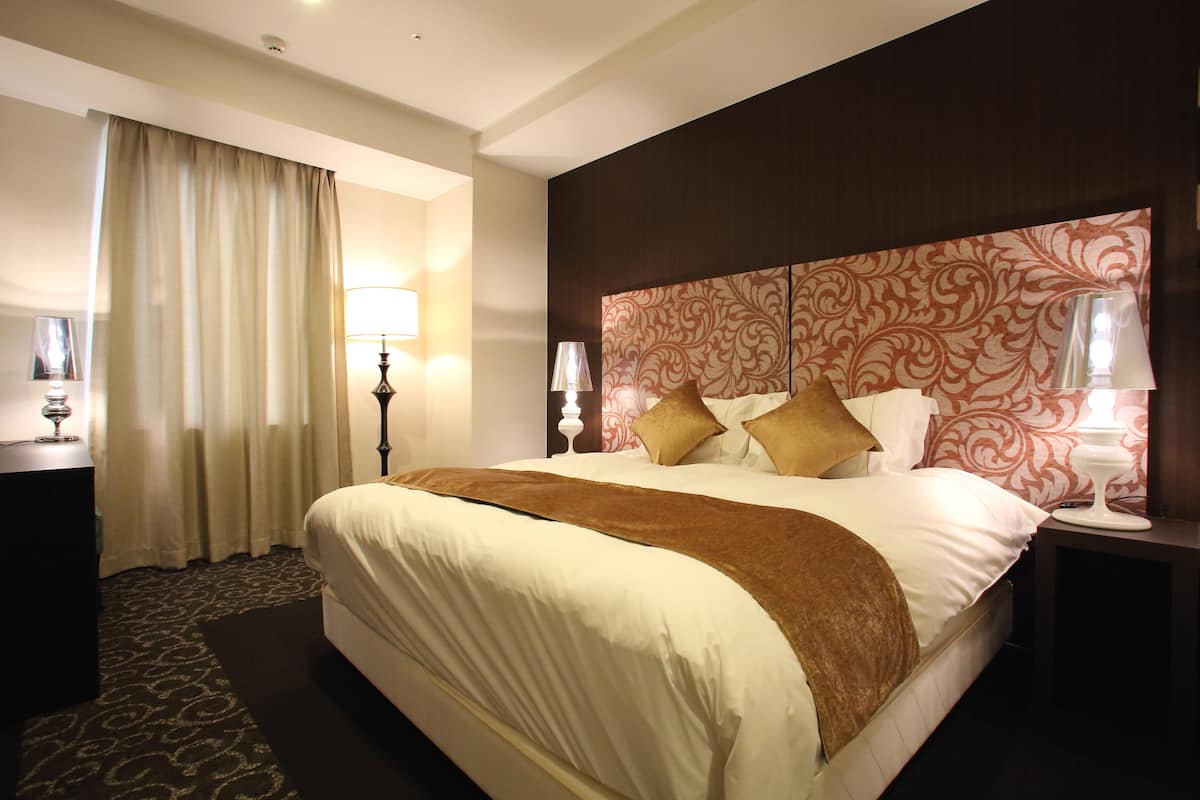 A hotel room with a large bed, two bedside lamps, a nightstand, a decorative headboard, and a window with curtains.