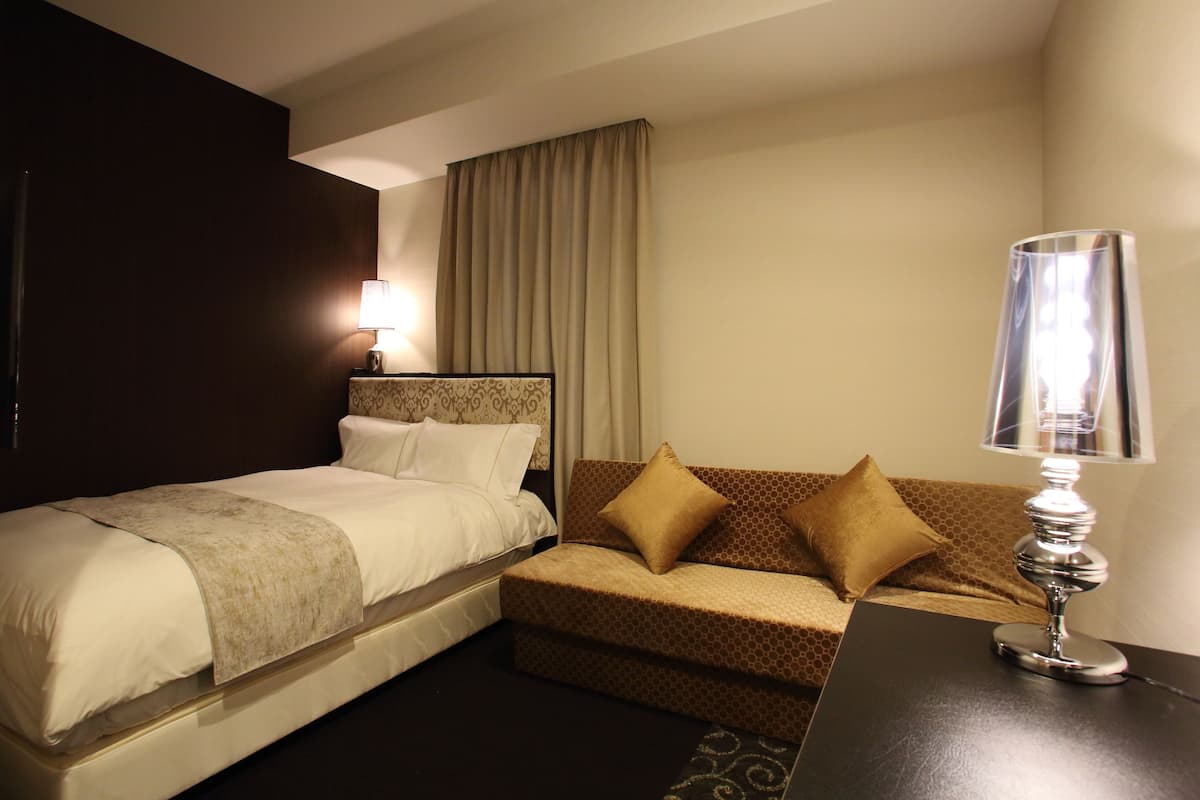 A hotel room with a bed, a sofa, a lamp, and a curtain.