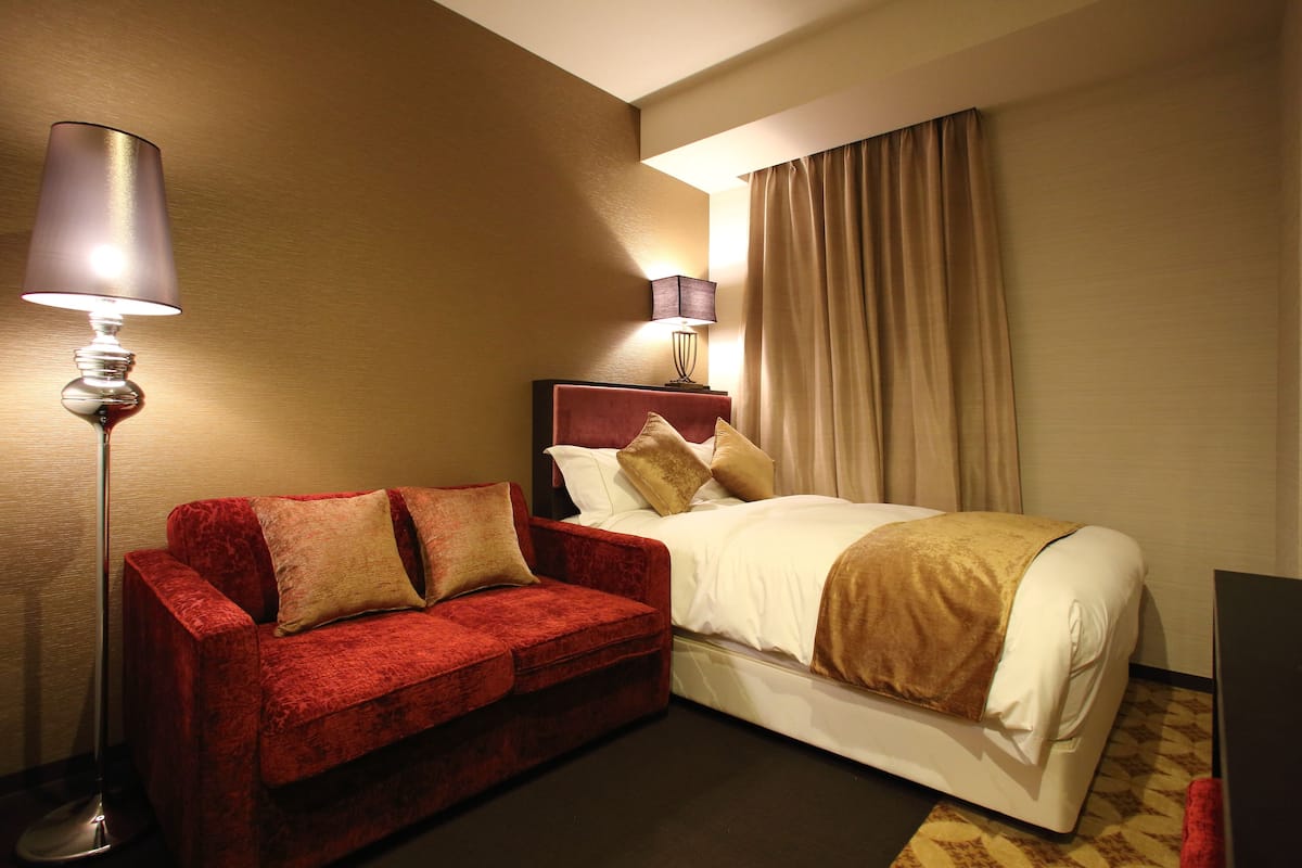 A hotel room with a red velvet sofa, a bed with white bedding, a floor lamp, and a wall-mounted lamp.