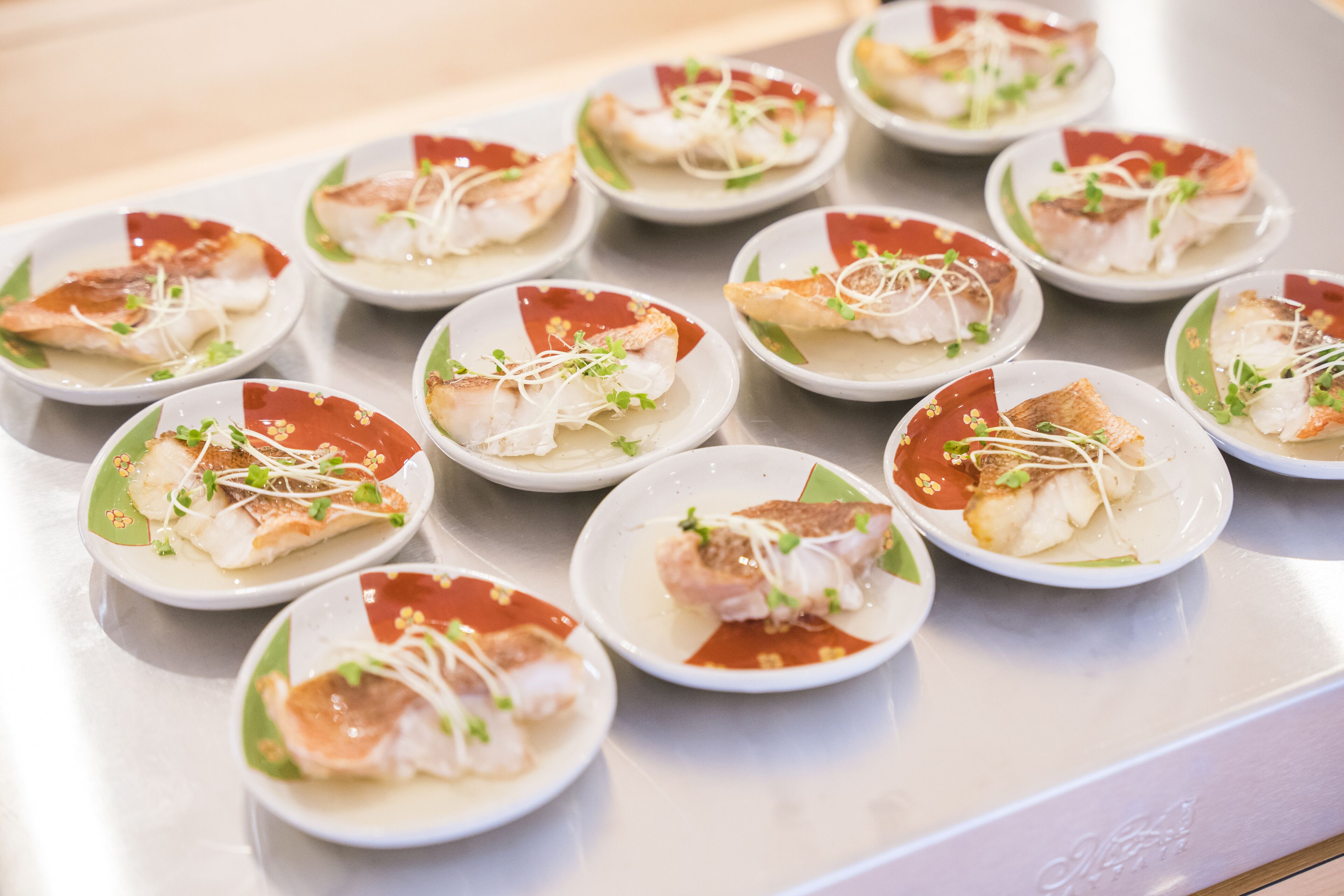Une table où sont disposées avec soin des assiettes remplies de nourriture, chacune contenant un morceau de poisson et agrémentée de jeunes pousses et d’une sauce rouge.