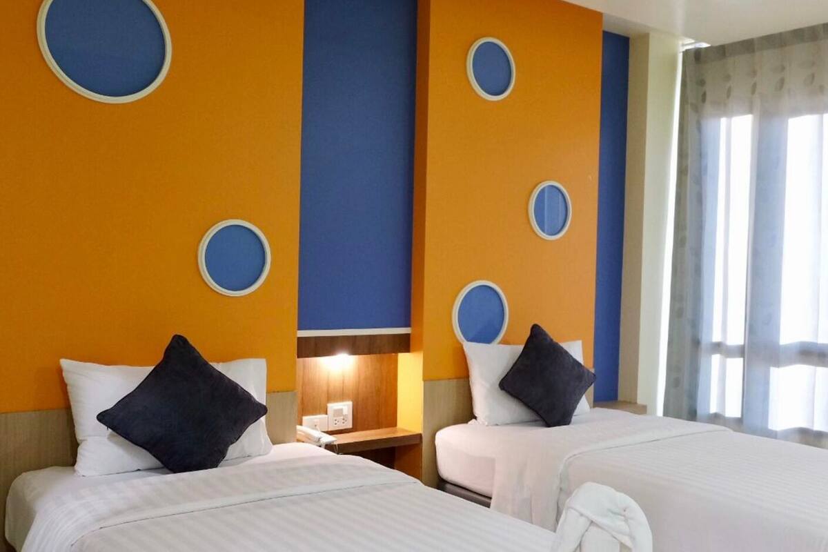 Ein Hotelzimmer mit zwei Betten, jeweils mit weißer Bettwäsche und dunklen Kissen. An den Wänden befinden sich runde Dekorationen und ein Fenster mit Vorhängen.