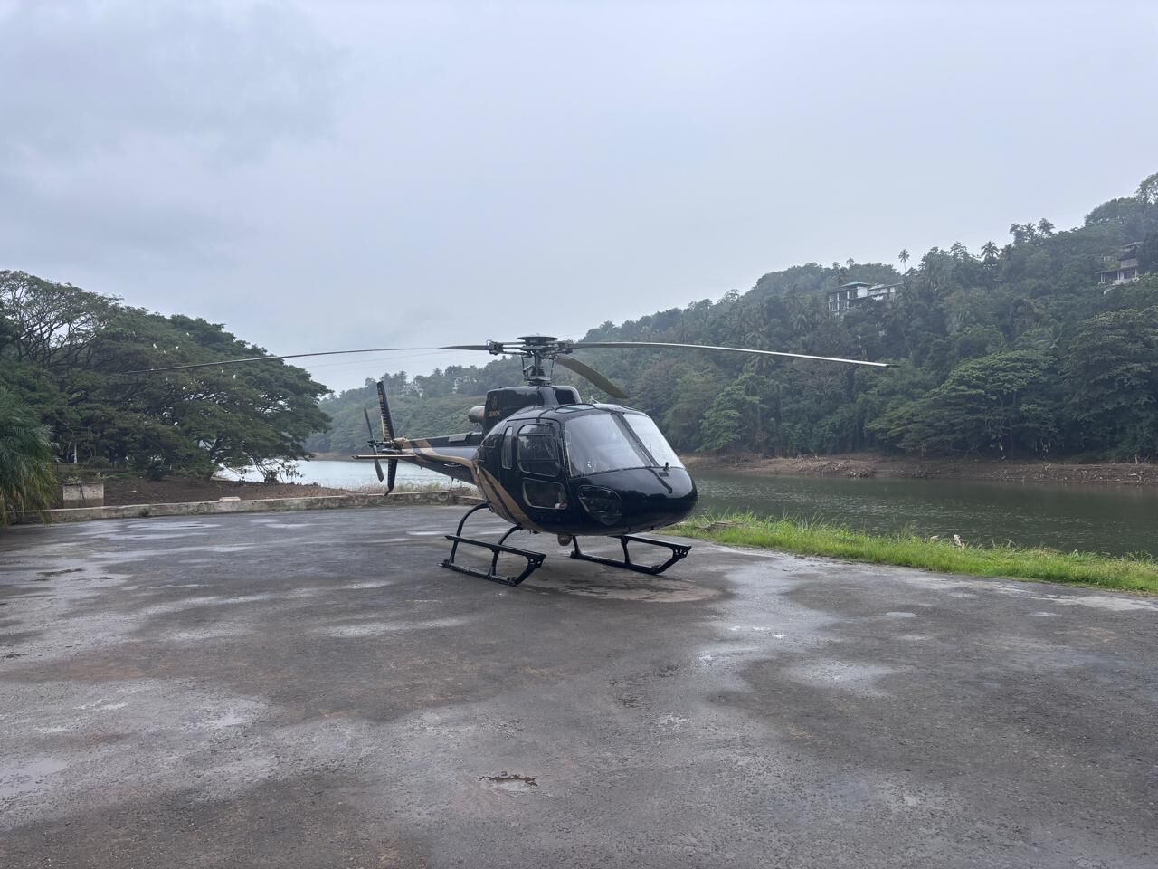 Ein Hubschrauber steht auf einer Betonfläche in der Nähe von Gewässer, mit Bäumen und Hügeln im Hintergrund.