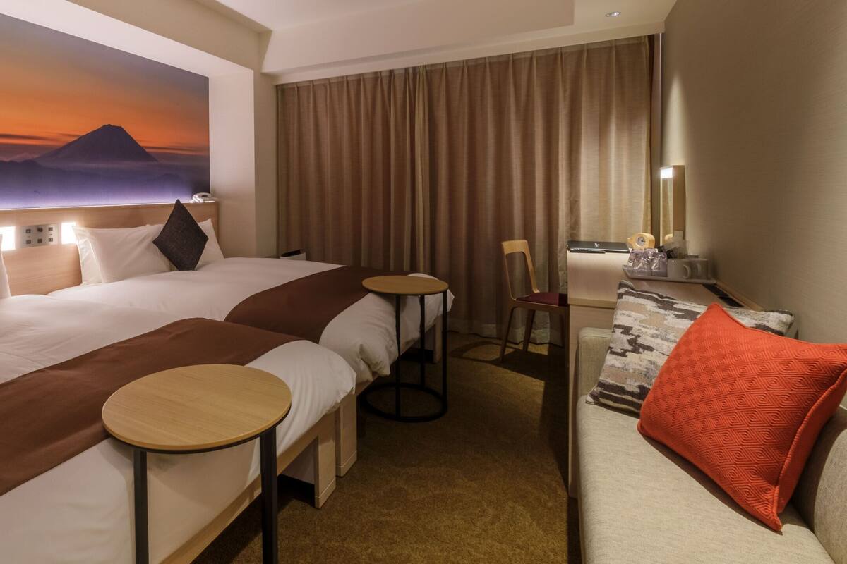 Ein Hotelzimmer mit zwei Betten, einem kleinen runden Tisch, einer Couch, einem großen Gemälde einer Berglandschaft und einem Vorhang.