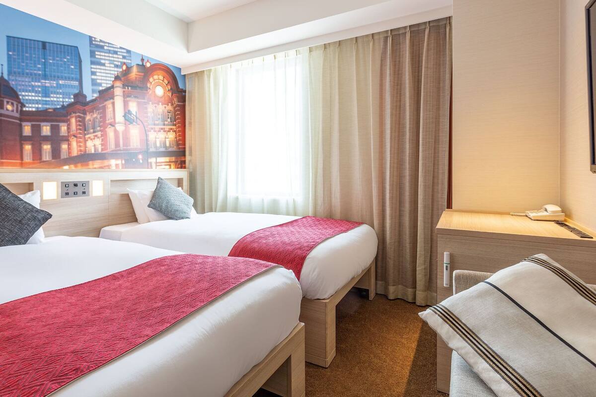 Ein Hotelzimmer mit zwei Betten, einem Schreibtisch, einem Sessel, einem Telefon und einem großen Fenster mit Vorhängen.