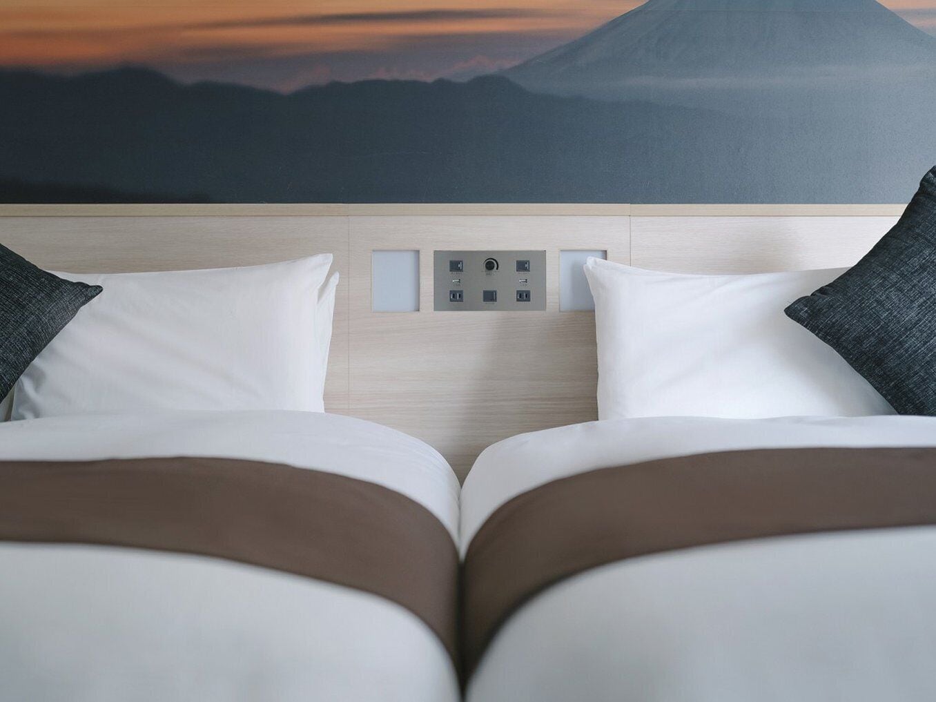 Ein Hotelzimmer mit einem Bett, ausgestattet mit weißer und brauner Bettwäsche, einer wandmontierten Steuerung und Blick auf einen Berg bei Sonnenuntergang.