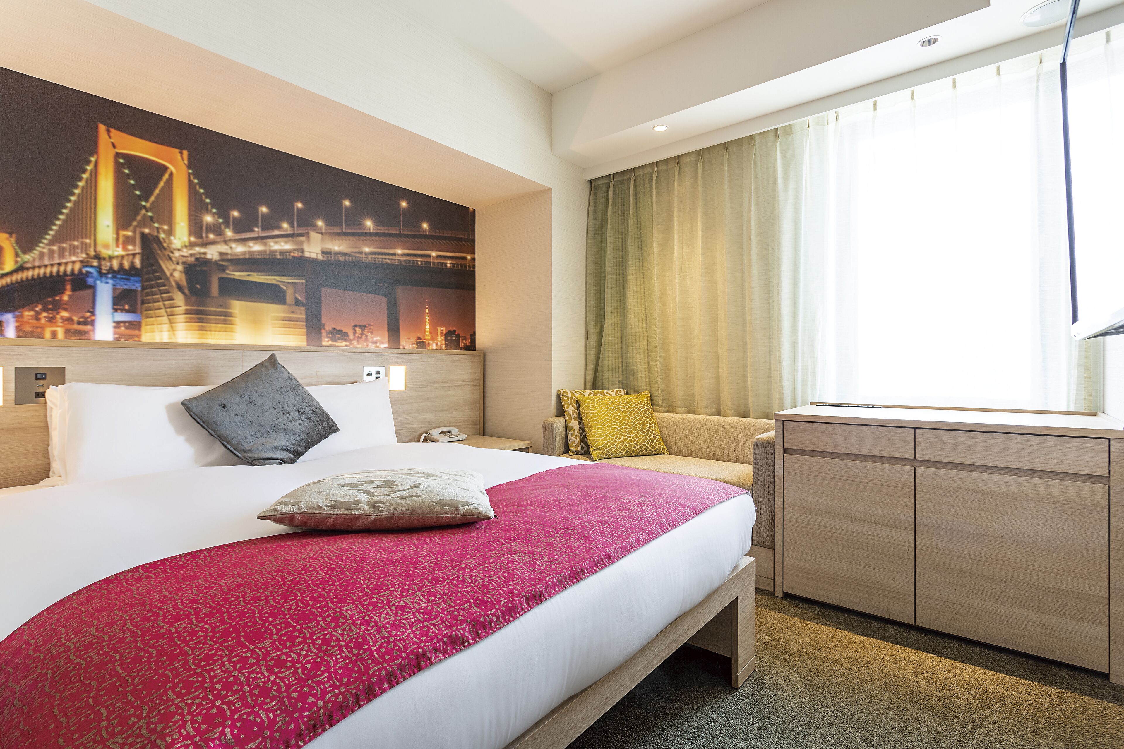 A hotel room with a large bed, a desk, a chair, a nightstand, and a cityscape mural on the wall.
