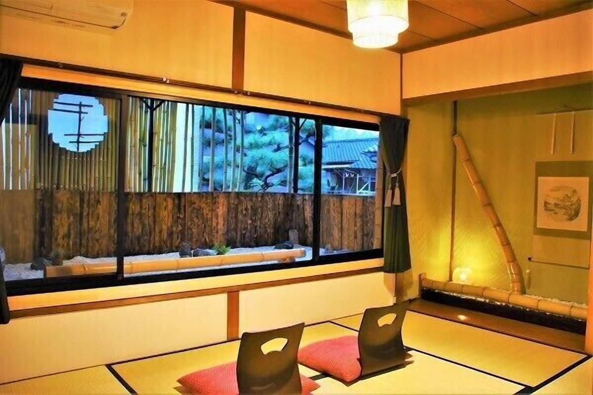 Una habitación japonesa tradicional con suelo de tatami, una mesa baja con cojines, un ventanal con vistas a árboles y un edificio, y un rastrillo de bambú.