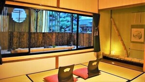 Una habitación japonesa tradicional con suelo de tatami, una mesa baja con cojines, un ventanal con vistas a árboles y un edificio, y un rastrillo de bambú.
